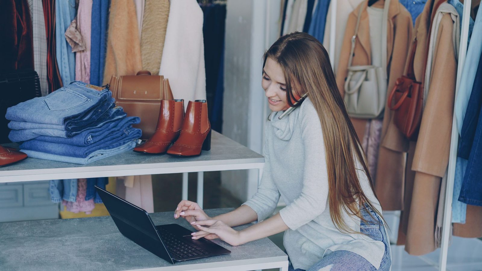 A shop owner on the phone types on a laptop at a counter in her clothing boutique.