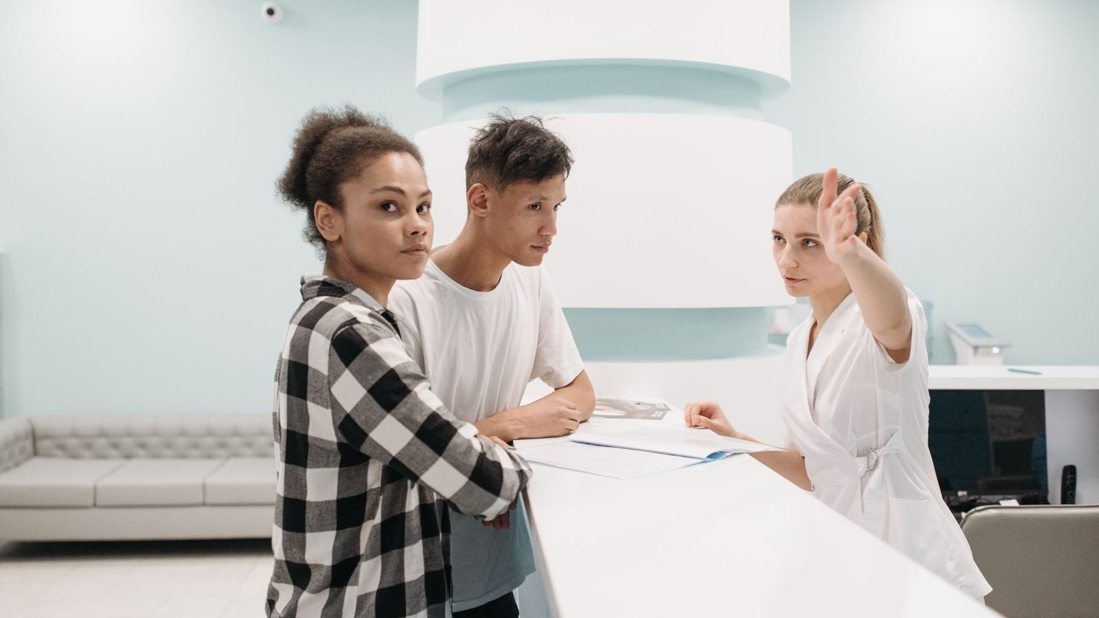 Two patients talk to a receptionist dressed in white at a clinic reception desk.