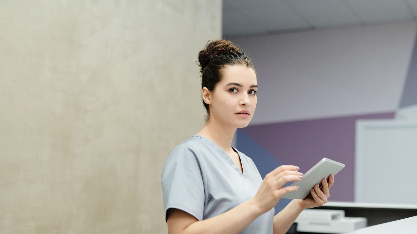 A receptionist in scrubs holds a tablet at a clinic reception desk.