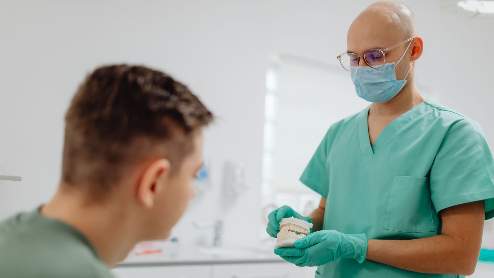 A dentist in green scrubs and a mask shows a dental model to a young patient.
