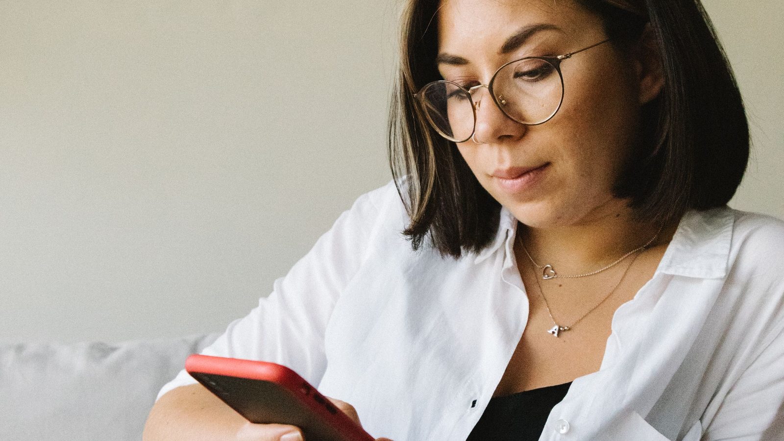 A woman in glasses and a white shirt sits on a sofa looking at her red phone.