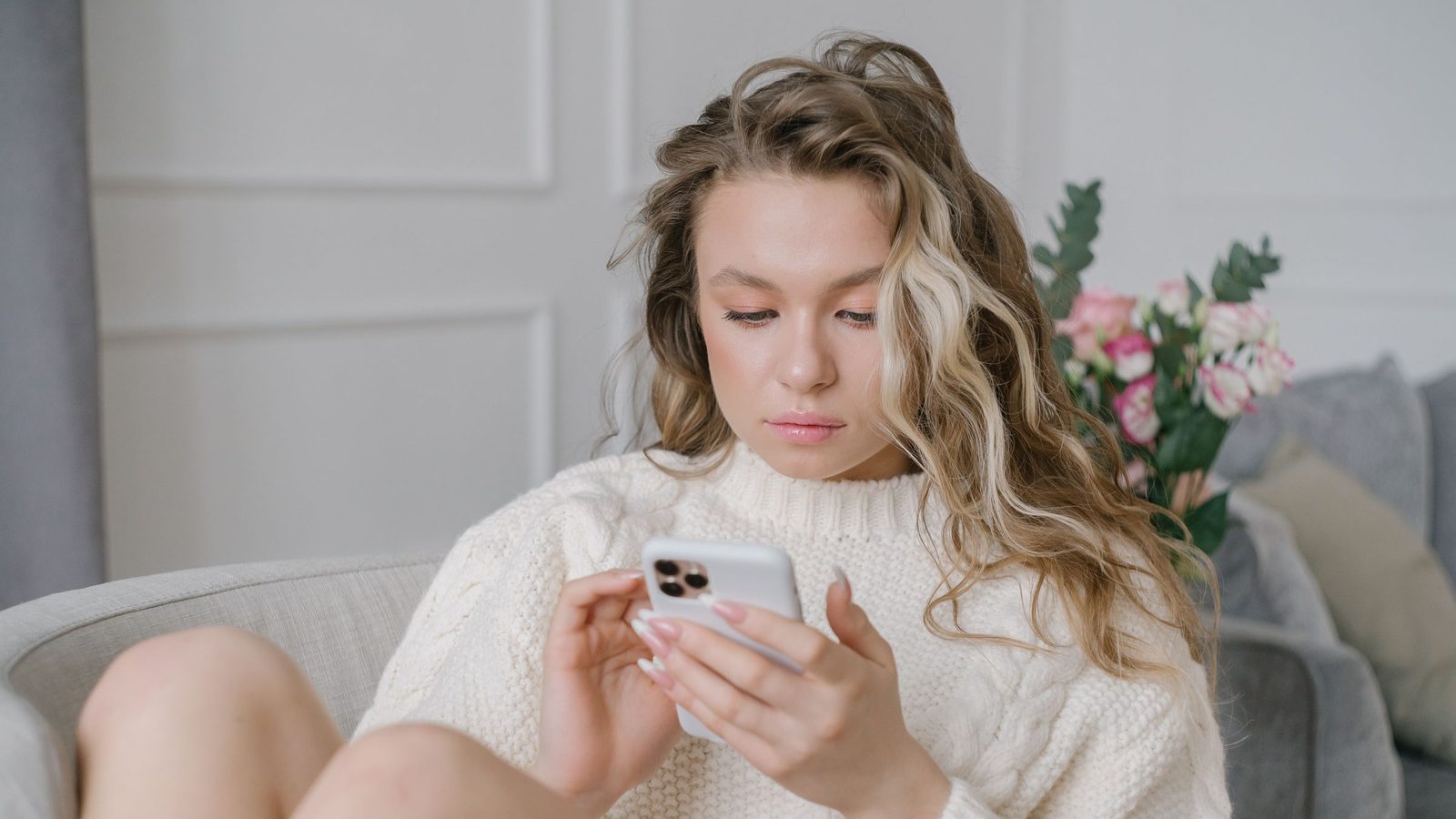 A woman in a cream jumper sits on a sofa typing on her phone.