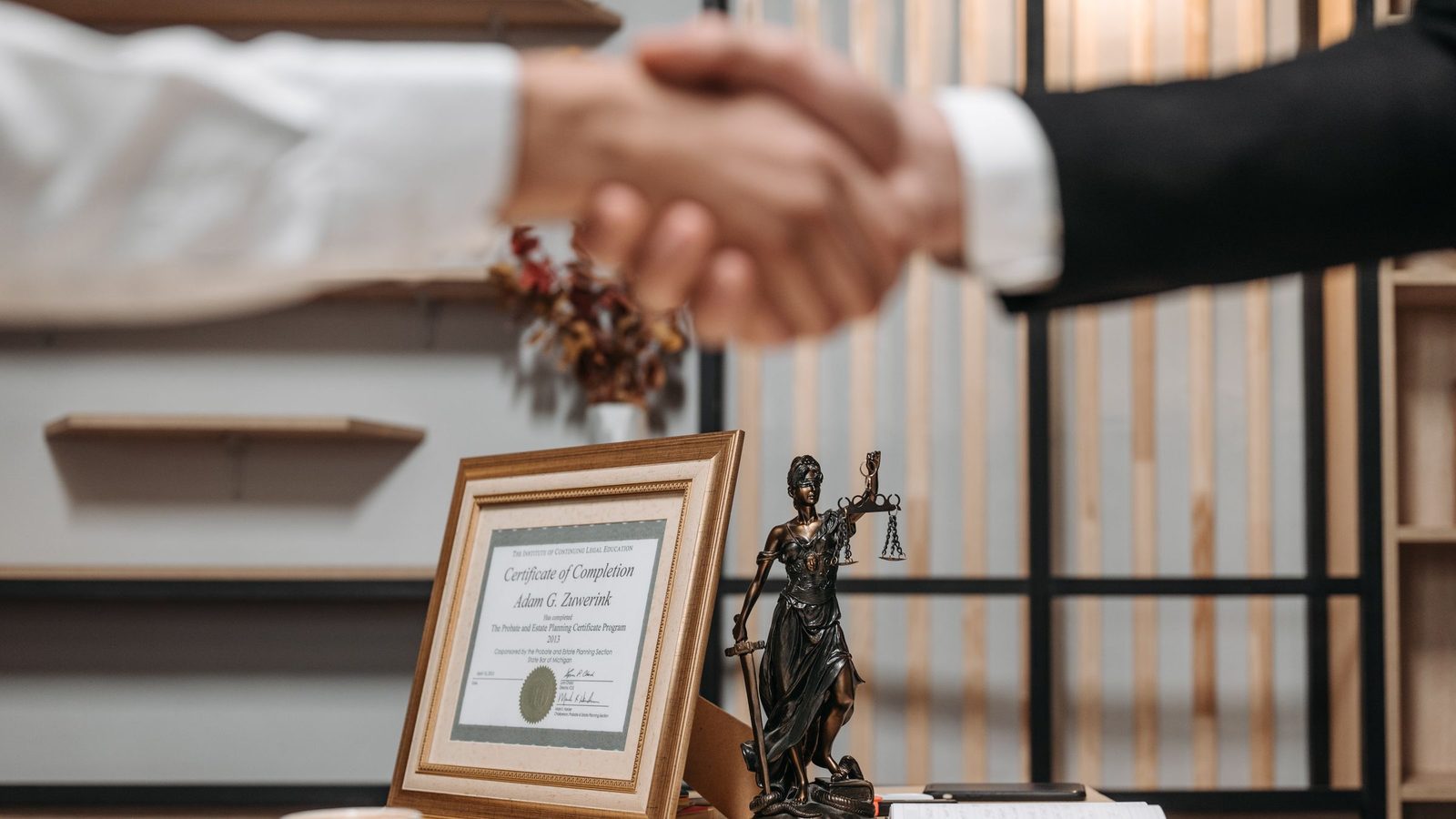 Two people shake hands across a desk beside a framed certificate and a small statue of Lady Justice.