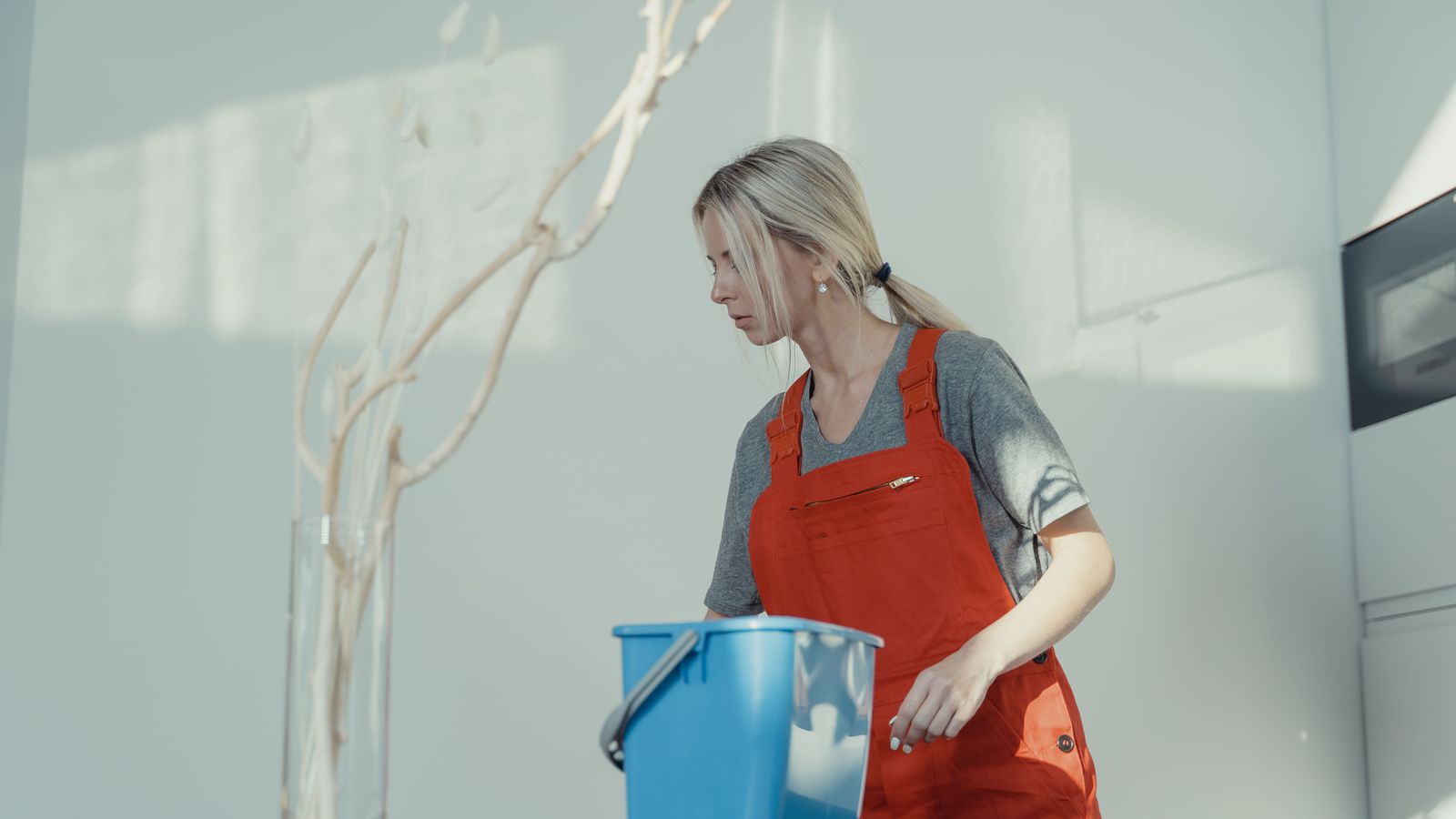 A cleaner in a red apron carries a blue bucket through a bright, modern office.