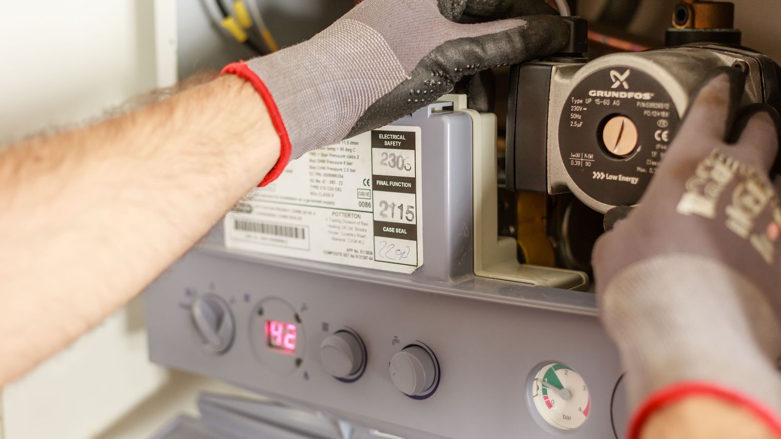 A heating engineer in work gloves fits the pump inside an open boiler with its display showing 42.