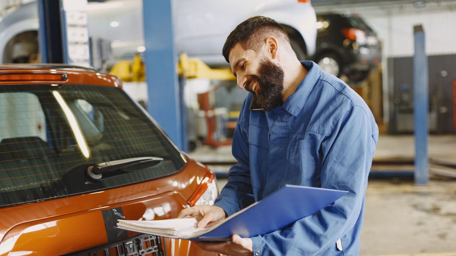 A smiling mechanic in blue overalls writes on a clipboard beside an orange car in a workshop.