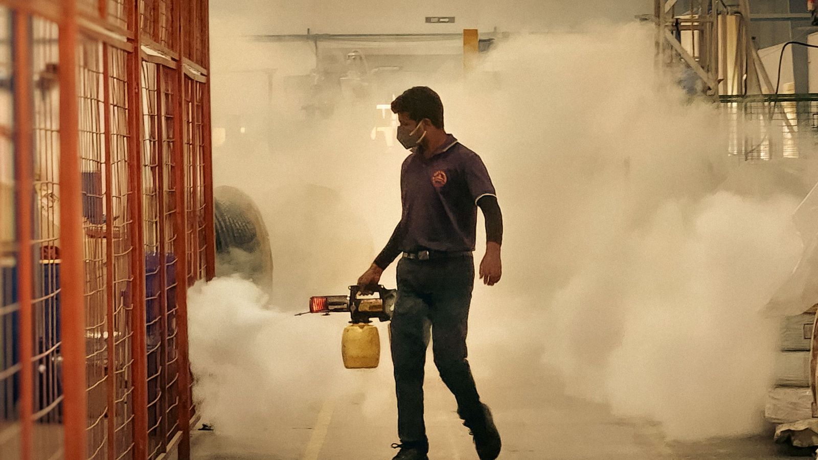 A pest control technician in a face mask fogs the aisle of a warehouse, surrounded by white mist.