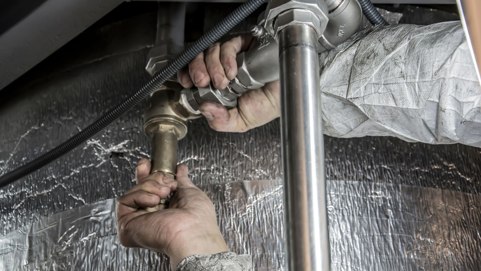 A plumber's hands tighten a brass fitting on a pipe run beside foil-lagged ductwork.