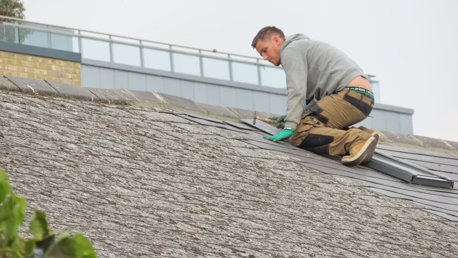 A roofer in work trousers and gloves kneels on a slate roof, fixing a slate near the ridge.