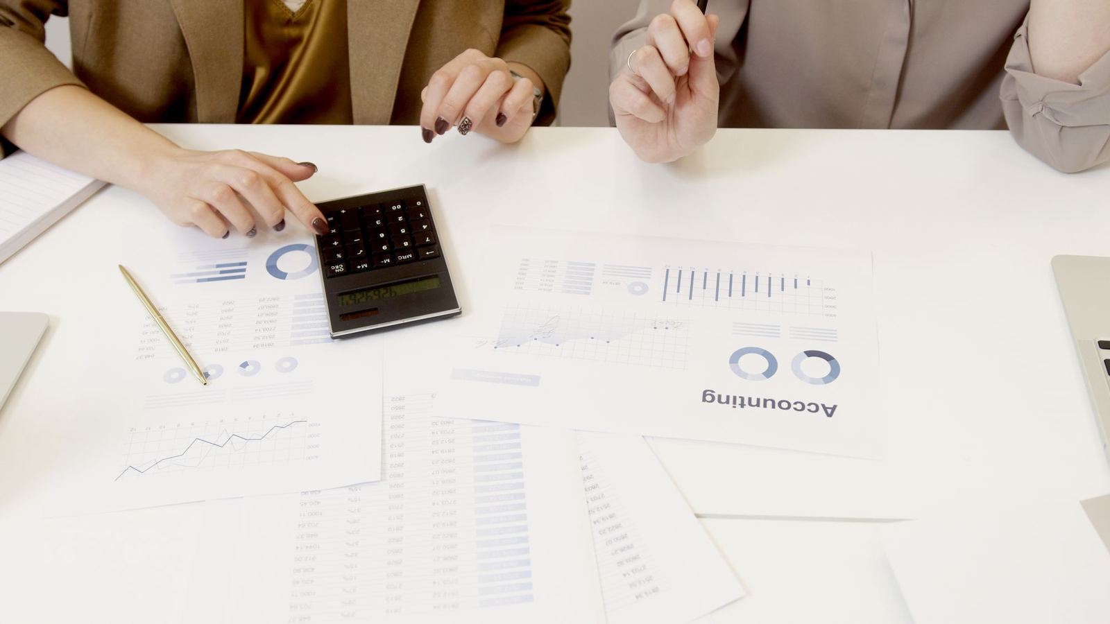 Two accountants go through printed charts and figures with a calculator at a white desk.