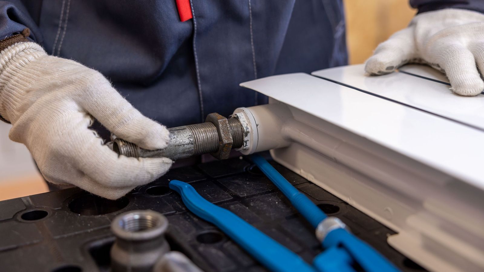 A plumber in work gloves tightens a threaded fitting into a white radiator, with blue pipe wrenches on the bench.