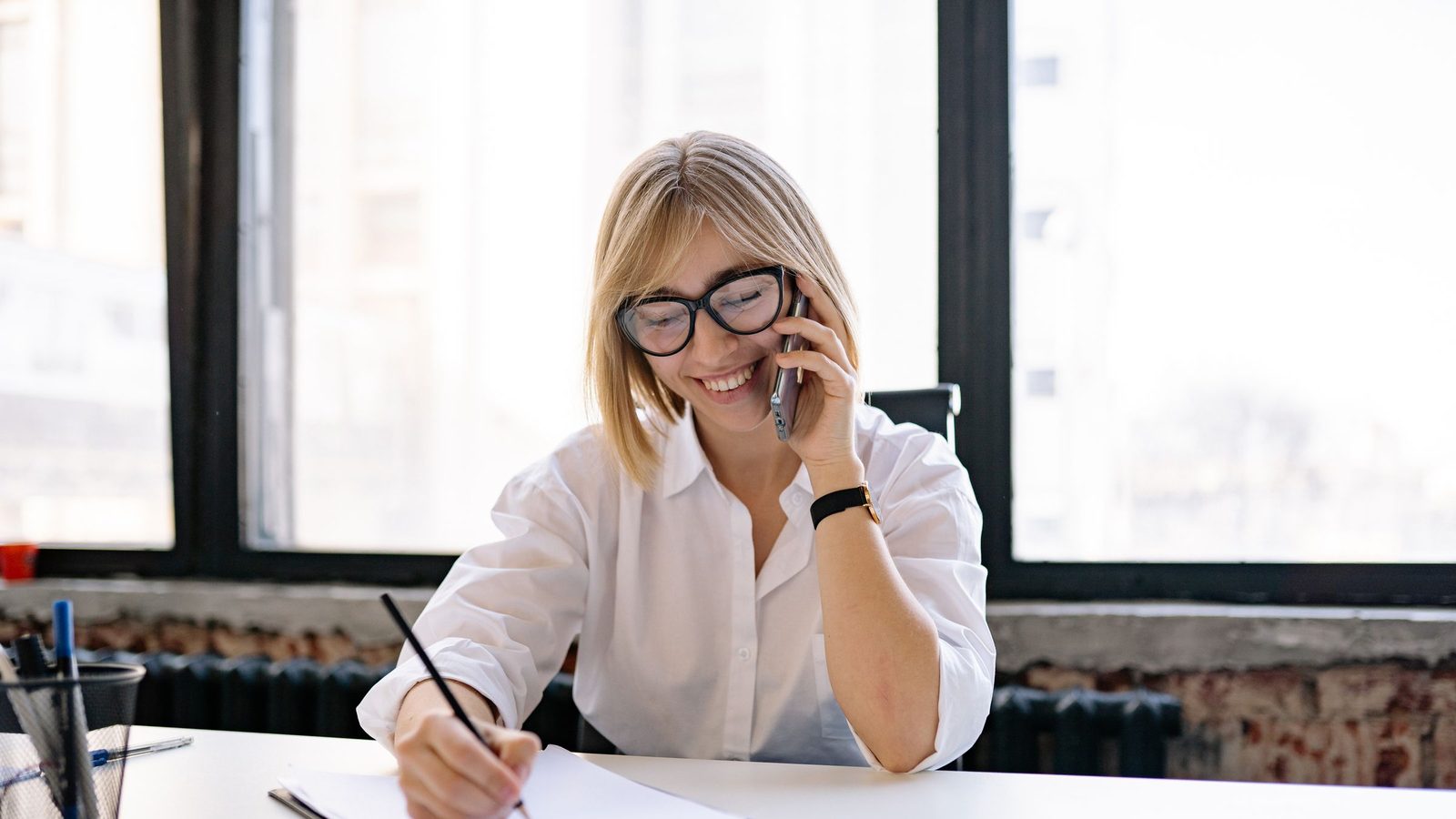 A smiling receptionist in glasses talks on the phone while writing notes at an office desk.