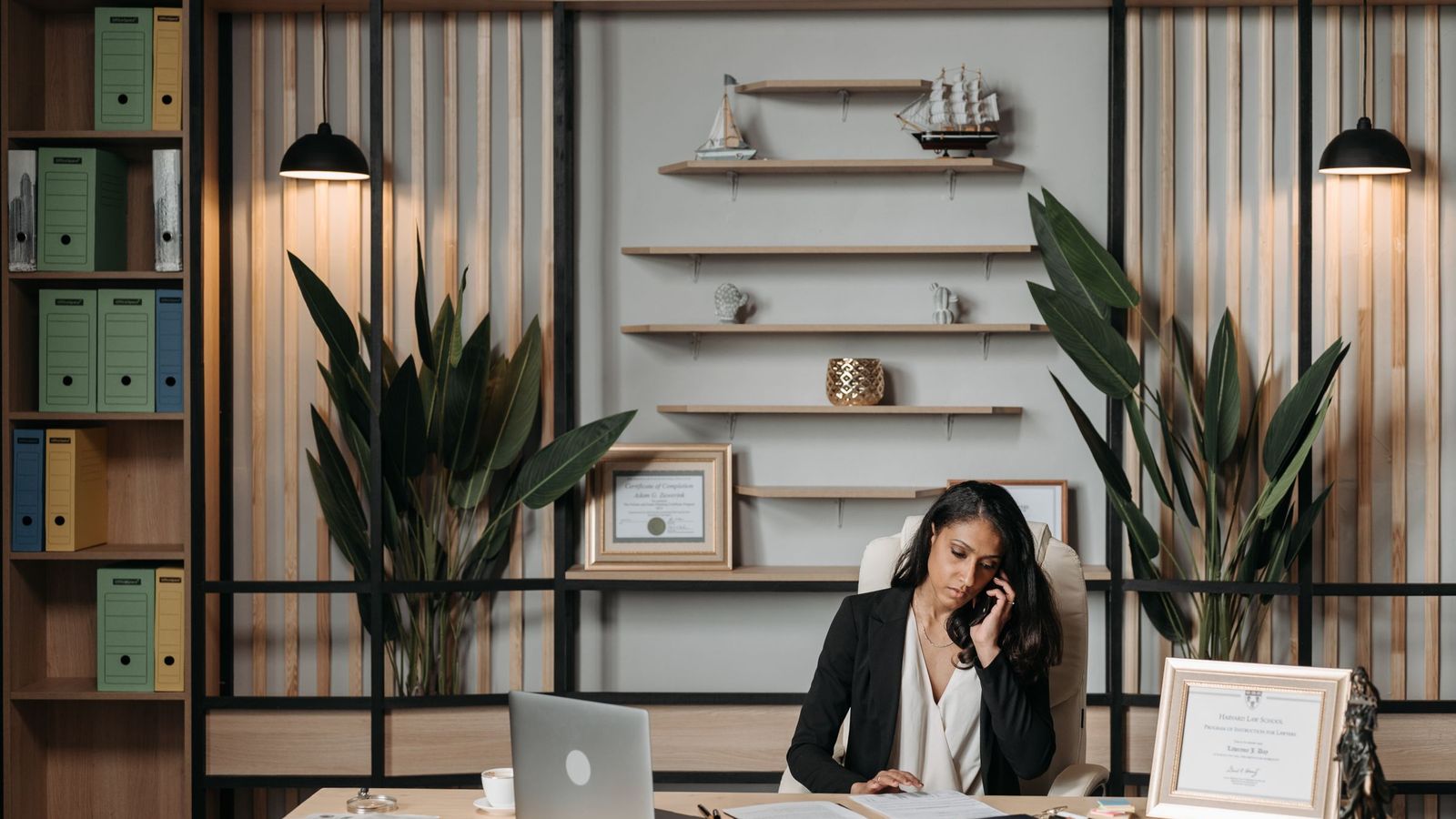 A woman takes a phone call at her desk with paperwork in front of her, in a bright office.