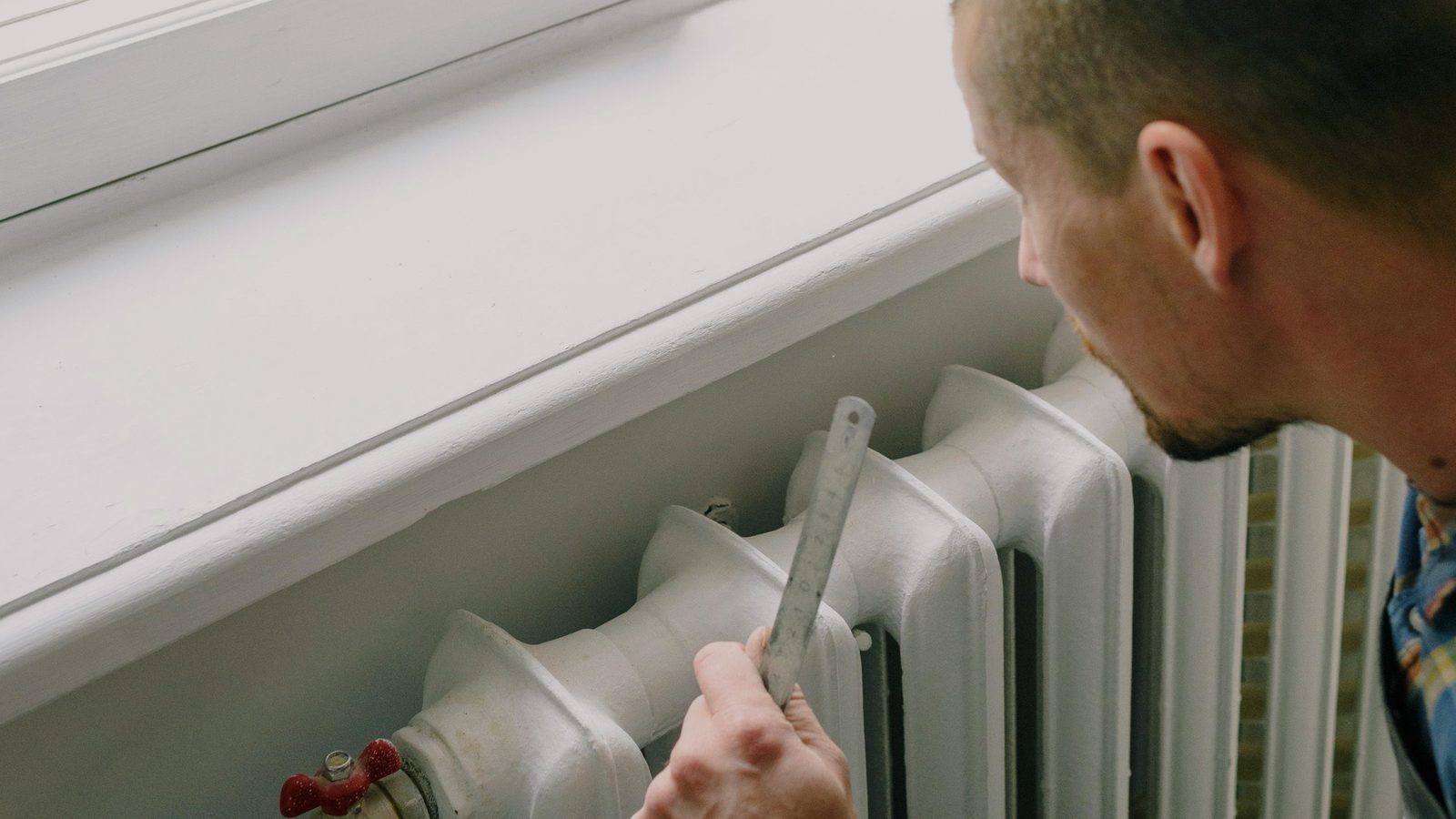 A man checks a white radiator under a window with a steel rule.