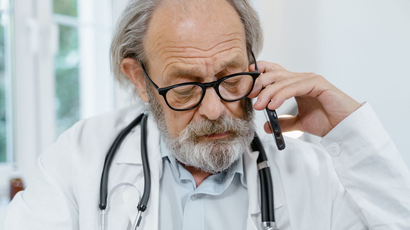 An older doctor in a white coat with a stethoscope talks on the phone.