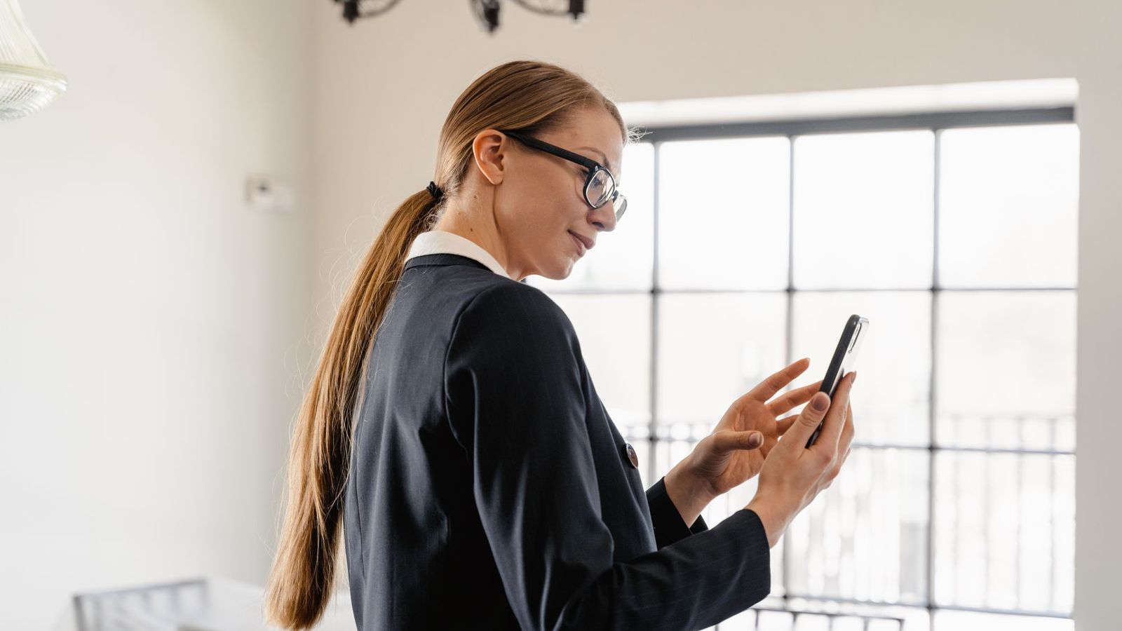 An estate agent in a dark suit and glasses checks her phone in a bright, empty room.