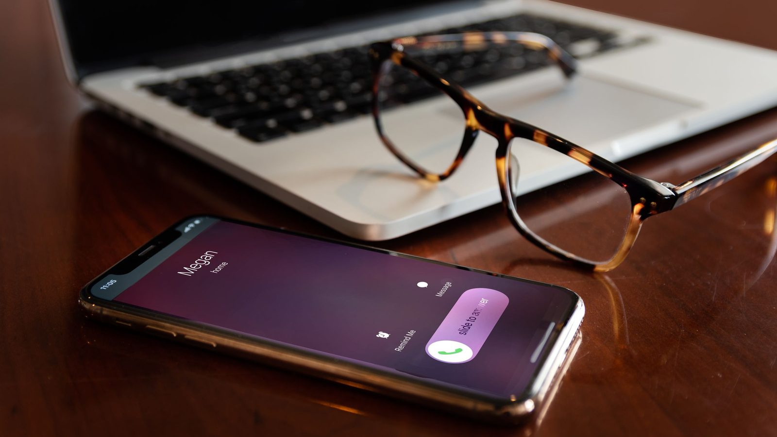 A smartphone showing an incoming call lies beside a laptop and a pair of glasses on a wooden desk.