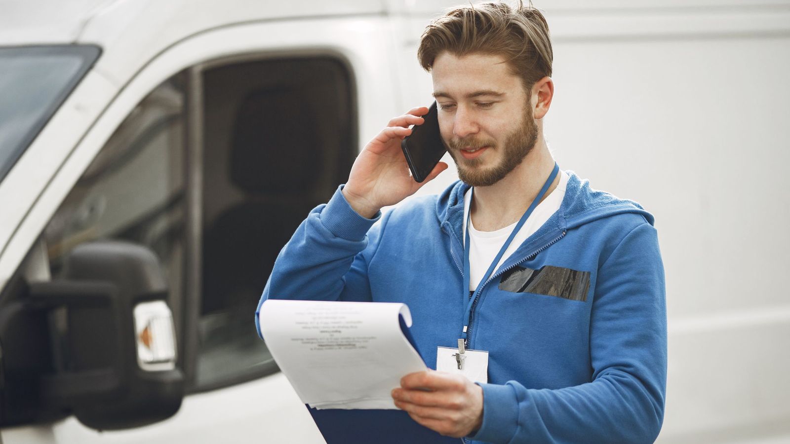 A man in a blue hoodie takes a phone call while holding a clipboard beside a white van.