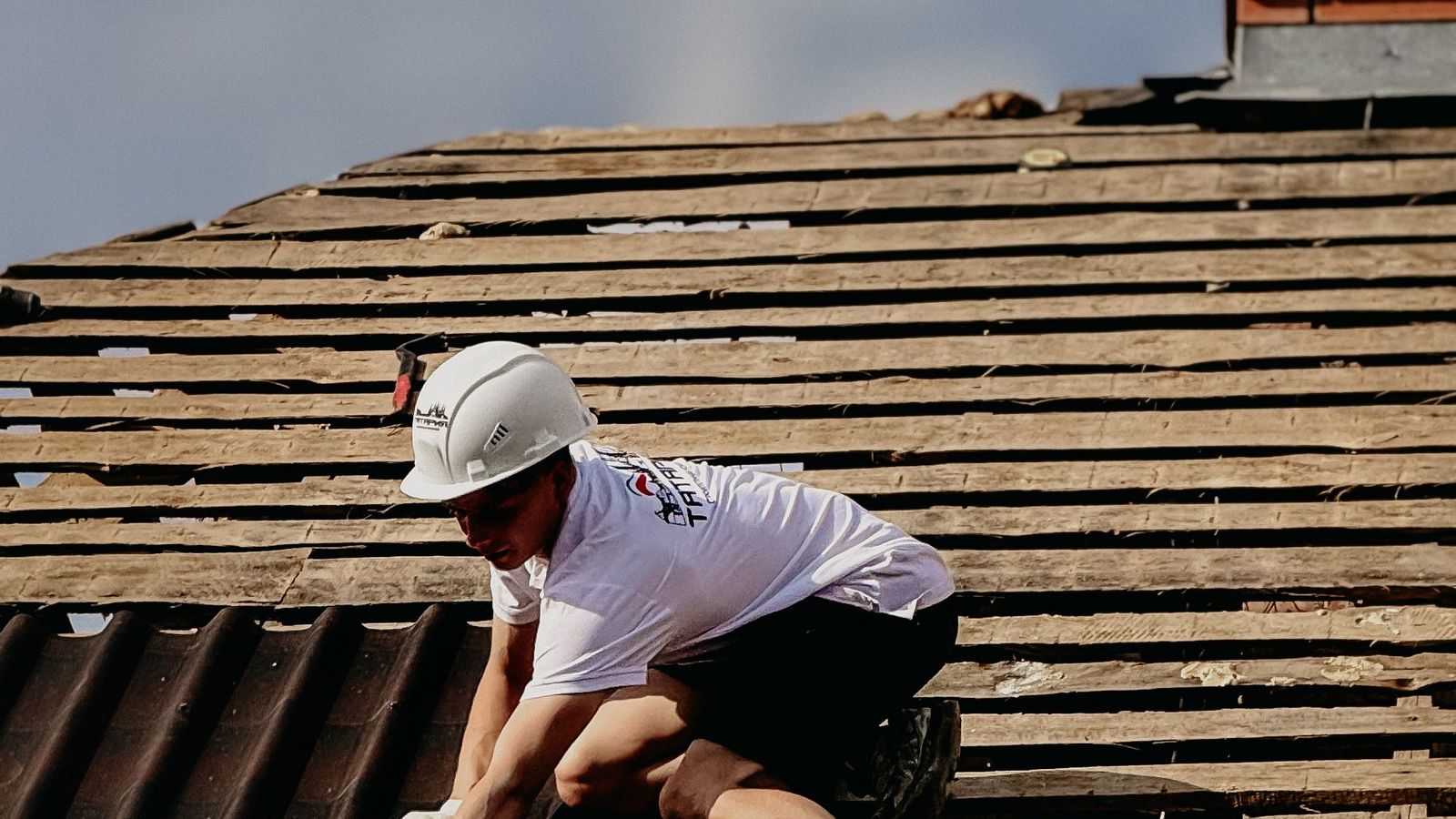 A roofer in a white hard hat works on a pitched roof stripped back to its battens.