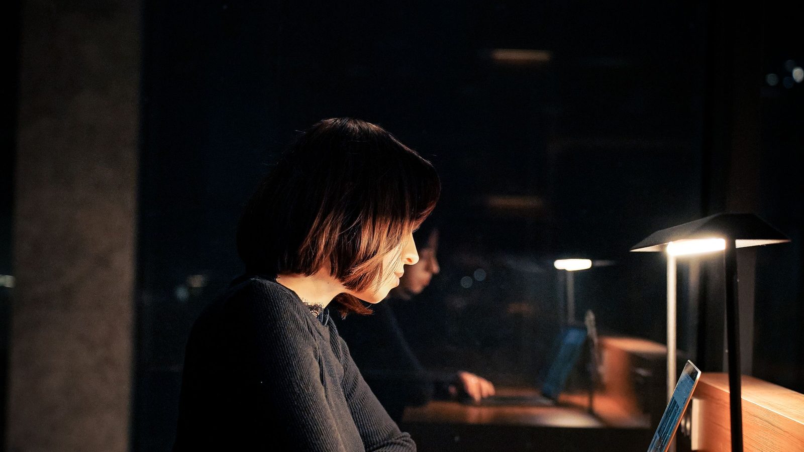 A woman works late at a desk in a dark office lit by desk lamps.