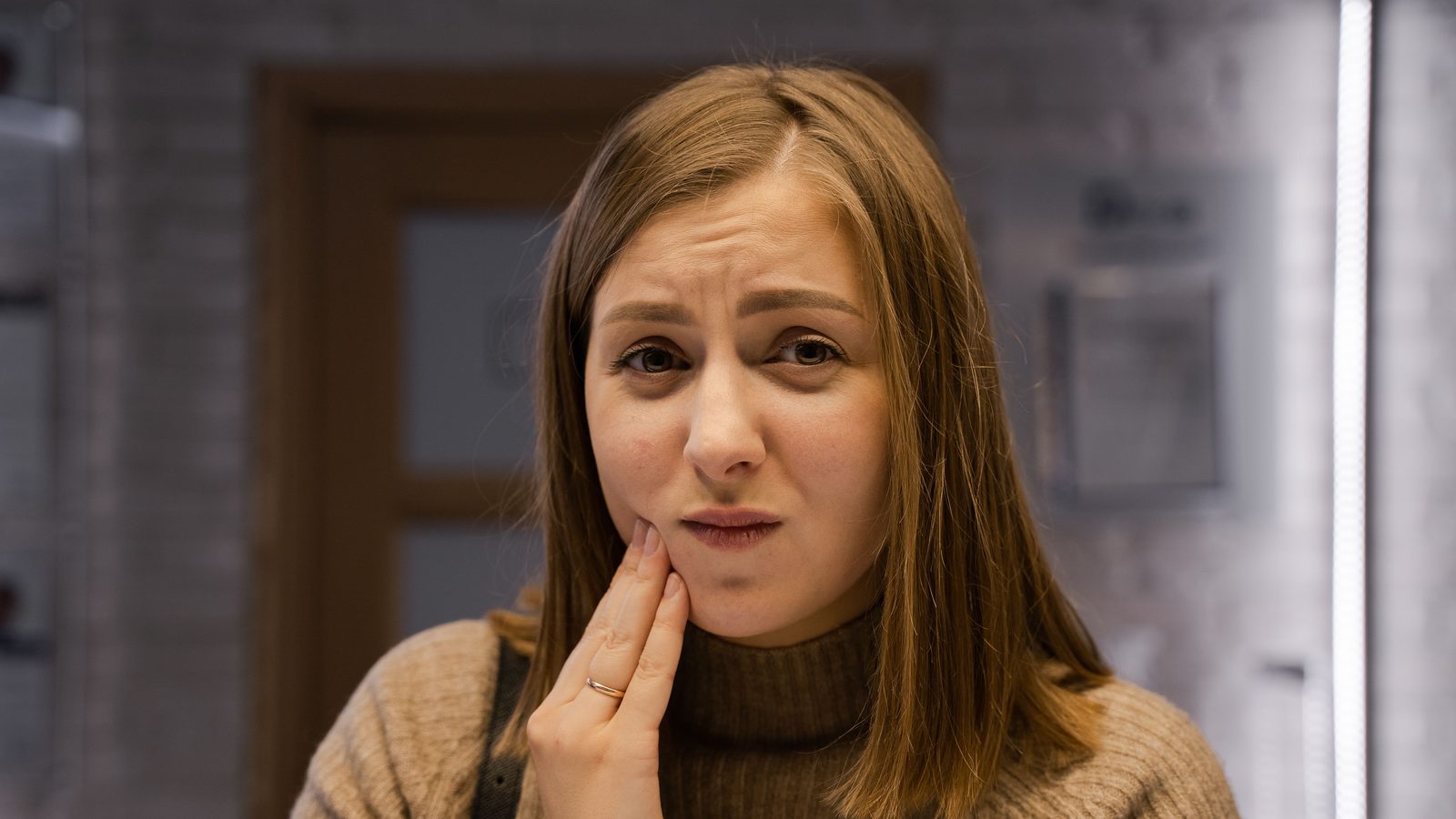 A woman in a brown jumper holds her cheek in pain at home.