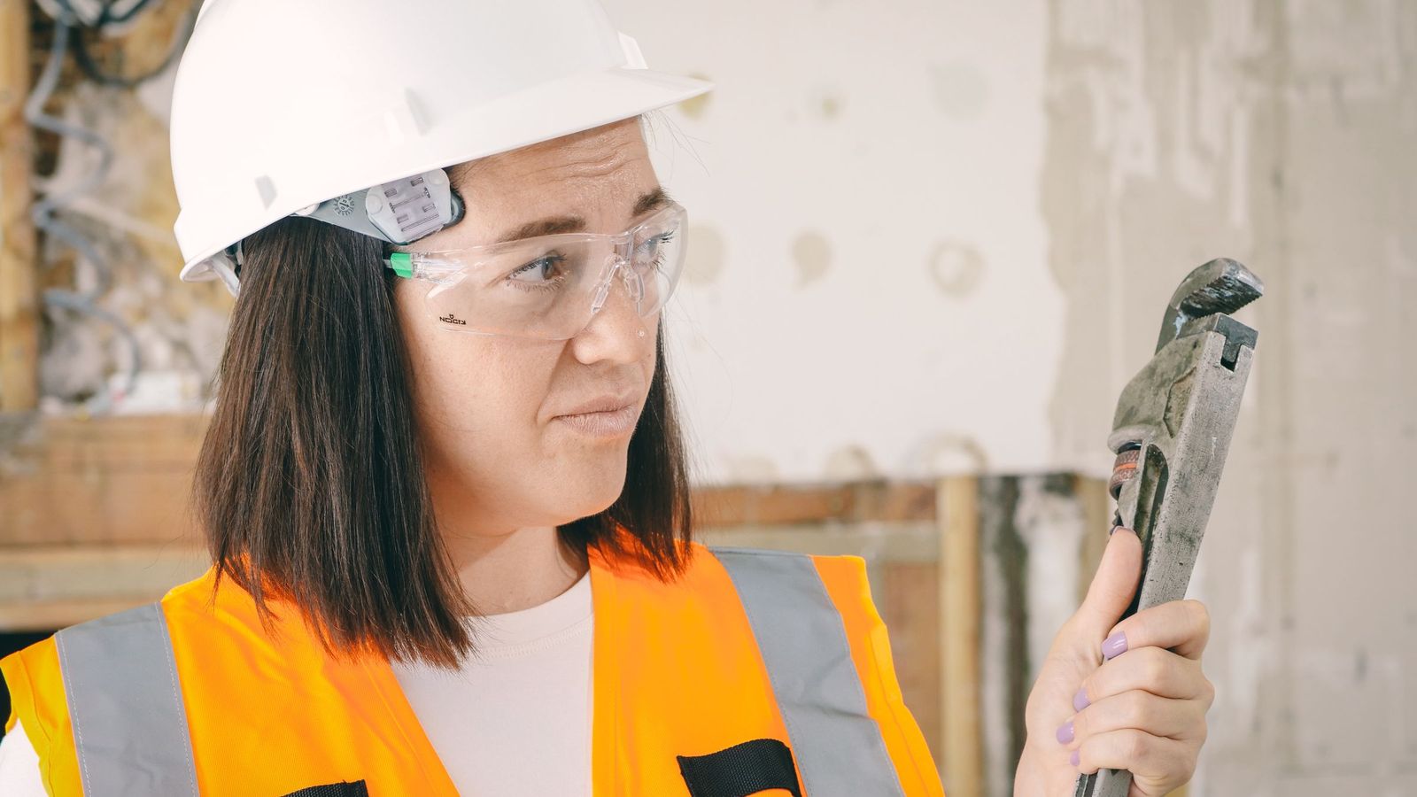 A plumber in protective gloves and glasses holds a pipe wrench indoors.