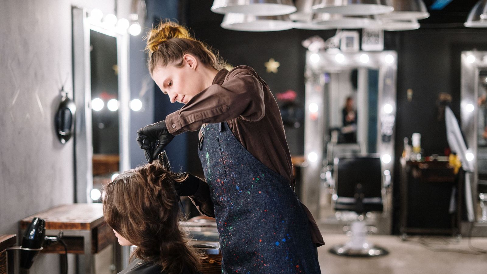 A stylist applies hair colour to a client's hair in a salon with mirrors and lights behind them.