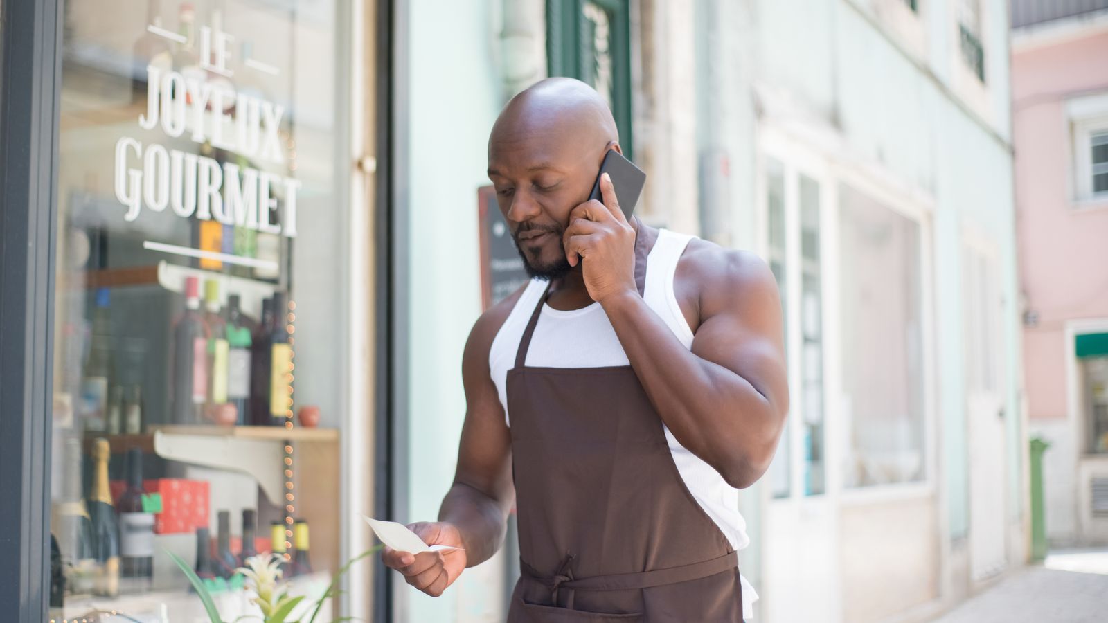 A shopkeeper in an apron talks on his phone outside his deli, checking a note.