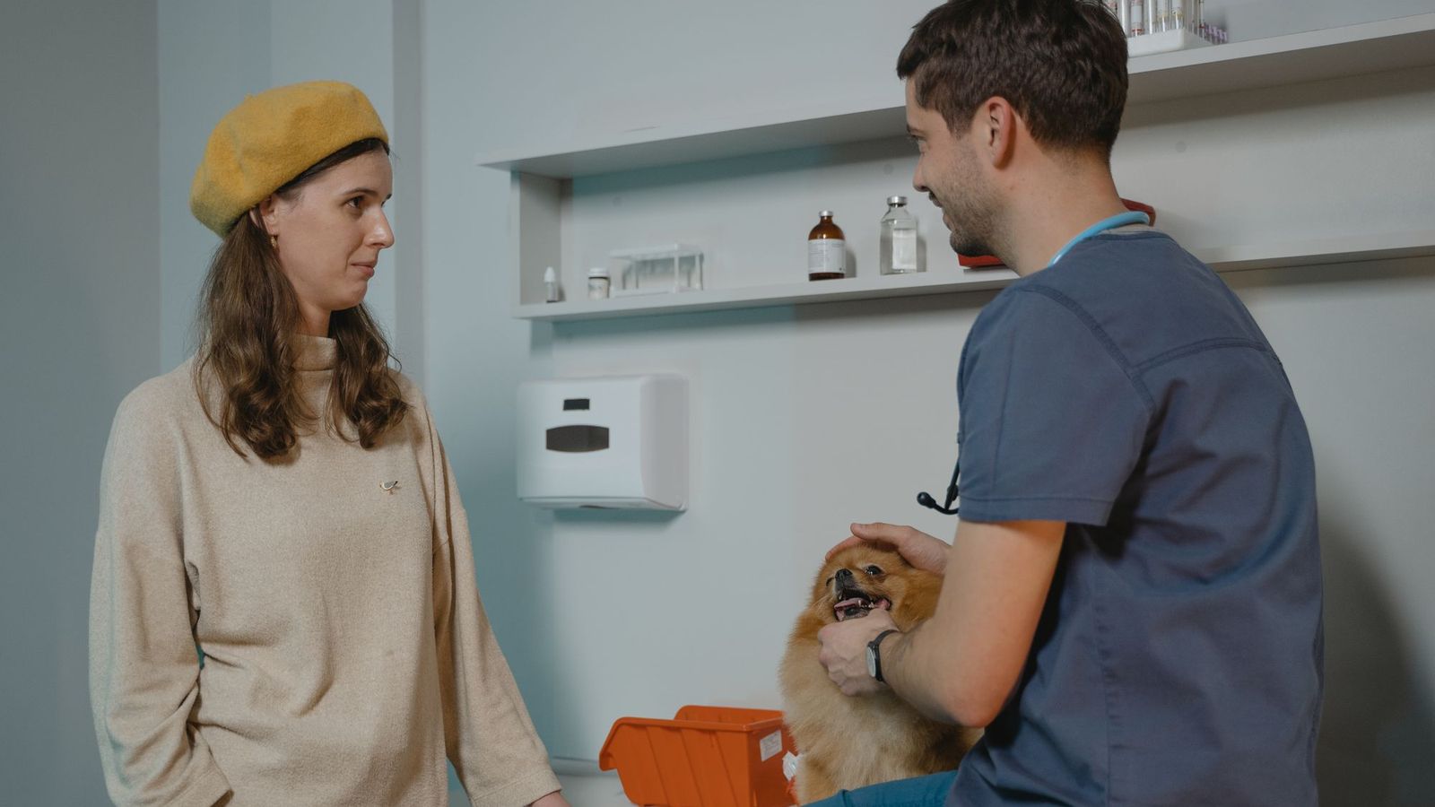 A vet in blue scrubs talks with a pet owner in a yellow beret who is holding her Pomeranian.