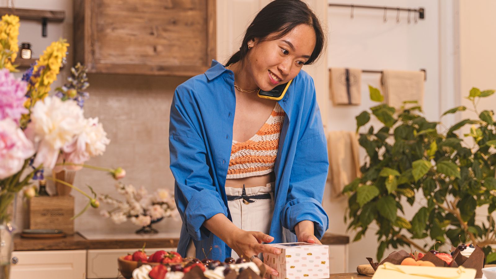 A baker on the phone boxes up an order at her kitchen counter.