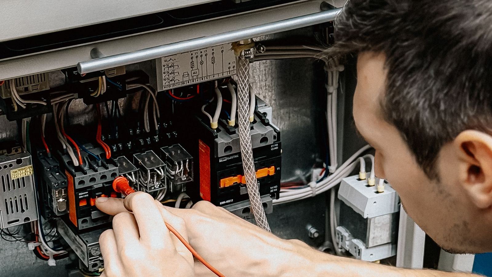 An electrician tests the wiring inside an open control panel with a multimeter.