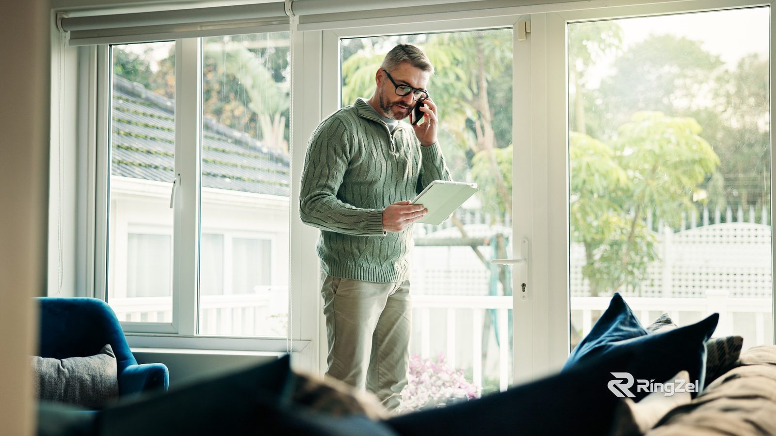 A man in a green jumper stands by a window on a phone call, reading from a tablet.