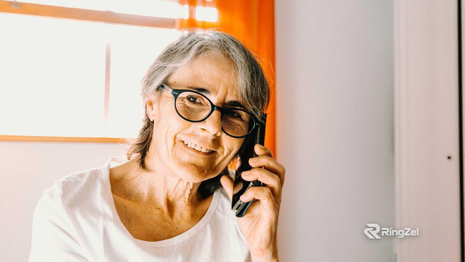 A smiling older woman with grey hair and round glasses talks on her mobile phone at home.
