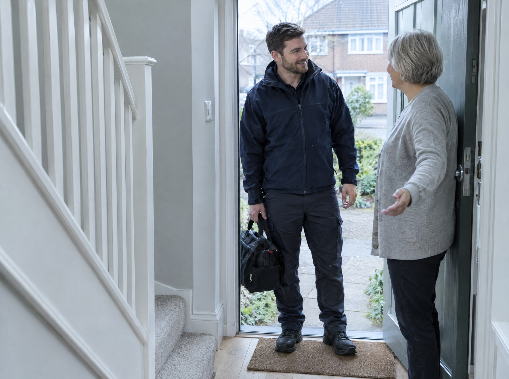 A heating engineer with a tool bag is welcomed at the front door by a homeowner.